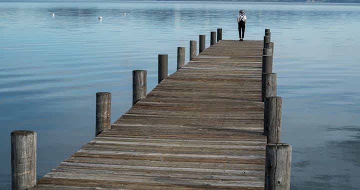 Distant traveler standing in solitude on long wooden pier with landscape of calm lake in highlands