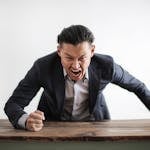Expressive angry businessman in formal suit looking at camera and screaming with madness while hitting desk with fist