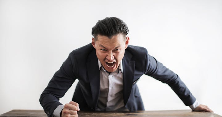 Expressive angry businessman in formal suit looking at camera and screaming with madness while hitting desk with fist